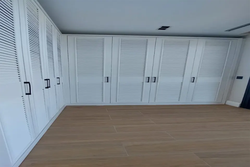 Ahşap parkeli odanın iki duvarını saran L biçimli panjur kapaklı dolap sistemi